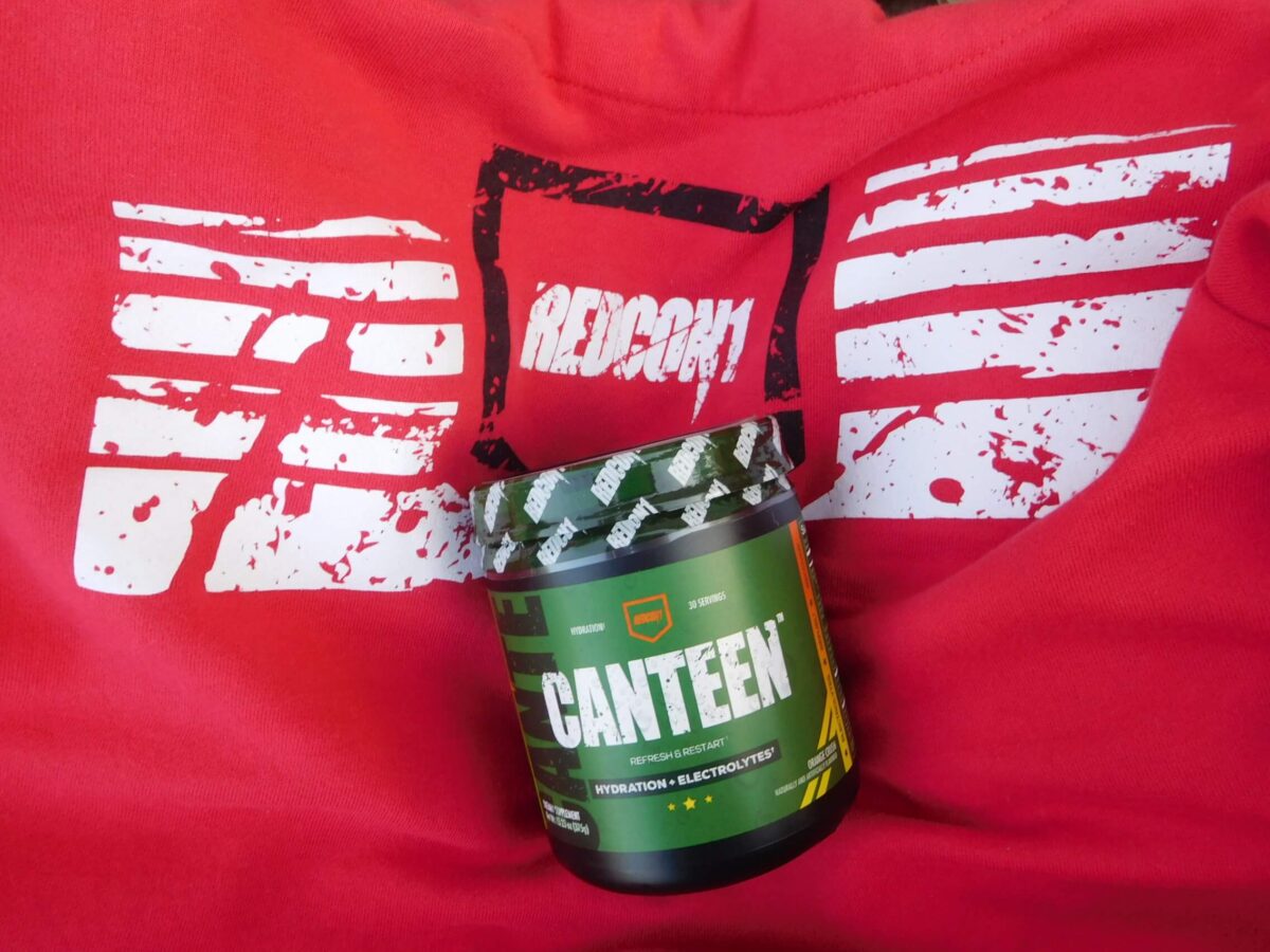 green tub of redcon1 canteen orange crush powder mix on top of red redcon1 hoodie with white and black logo