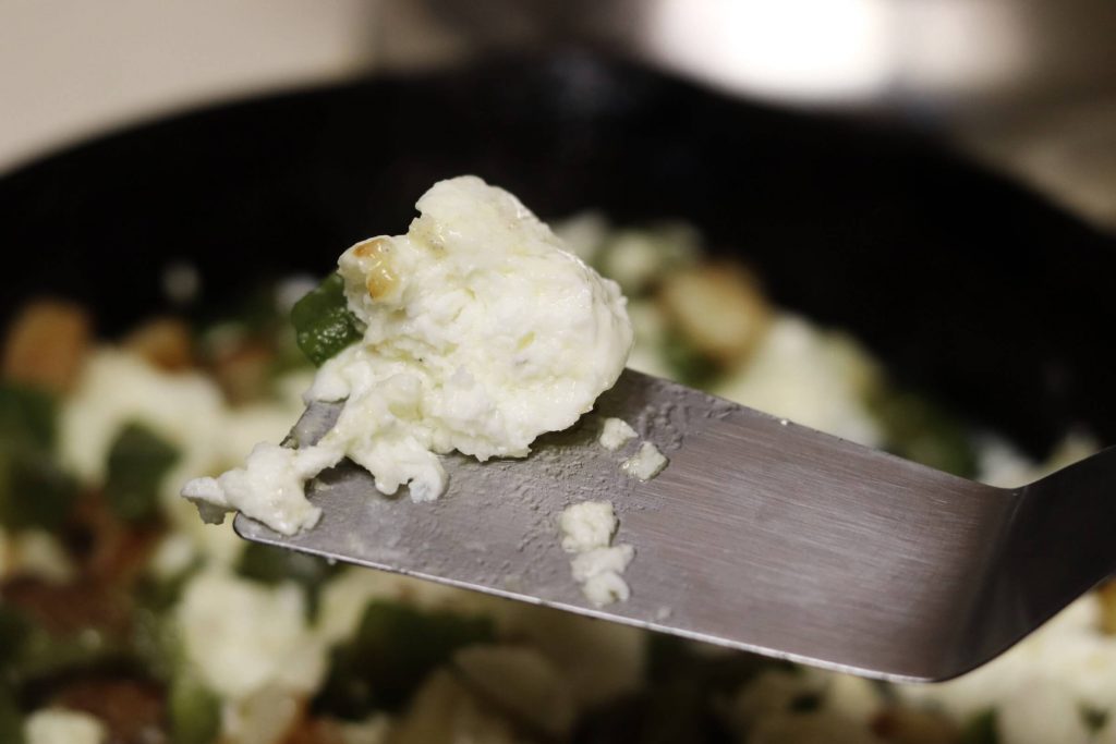 closeup of scrambled egg white chunk on spatula with blurry image of skillet with scrambled eggs, green peppers, potatoes and mushrooms in the background
