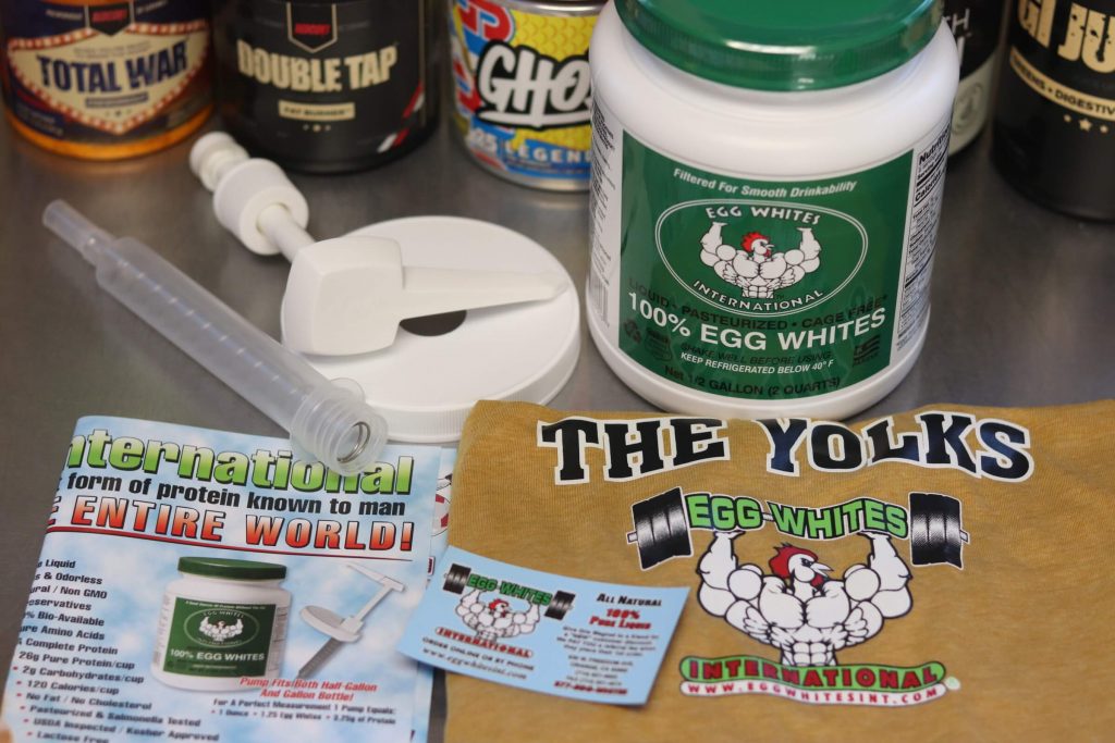 half gallon tub of egg whites international liquid egg whites with green label and cartoon muscular chicken next to disassembled pump with folded mustard yellow egg whites international shirt and folded pamphlet for egg whites international