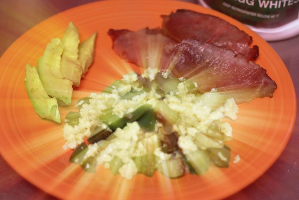 large orange plate with four slices of avocado, 3 slices of bacon, and pile of scrambled egg whites with potatoes, green peppers and mushrooms with corner of tub of egg whites international liquid egg whites in top right corner reading 100% egg with a filter on the picture that makes it look like a sunburst is coming out of the scrambled eggs