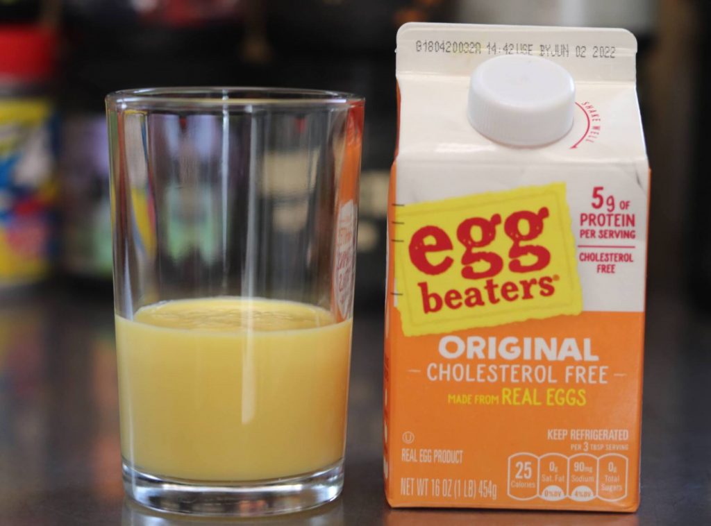small orange and white carton of egg beaters cholesterol free liquid egg whites next to a glass half filled with orangish liquid egg whites