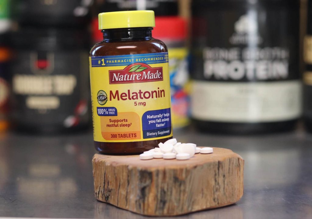 wood block with pile of white pills in front of a bottle reading nature made melatonin 5 mg supports restful sleep naturally helps you fall asleep faster dietary supplement 300 tablets