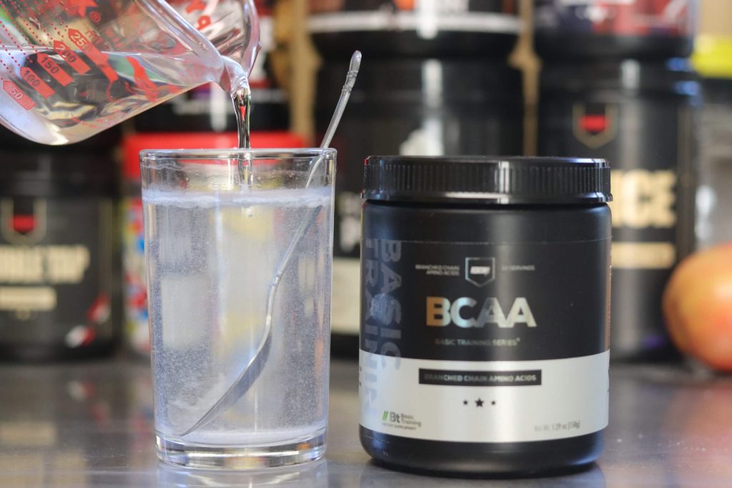 tub of redcon1 bcaa with black and silver label next to glass containing slightly cloudy mixture of bcaa powder and water with measuring cup pouring in more water