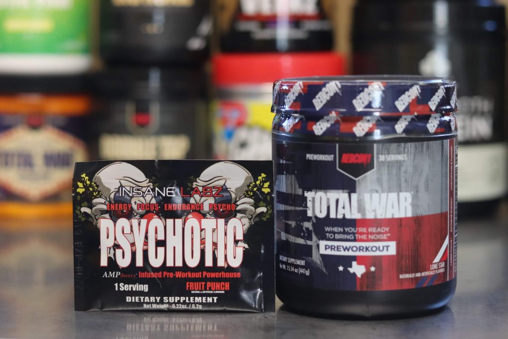 sealed tub of redcon1 total war pre-workout lone star flavor next to packet of insane labz psychotic pre-workout fruit punch flavor with two evil clown drawings