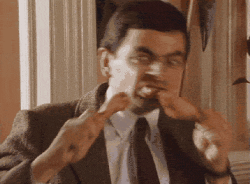 gif of mr bean man with short hair and suit and tie quickly biting chicken drummies in each hand while aggressively raising eyebrows