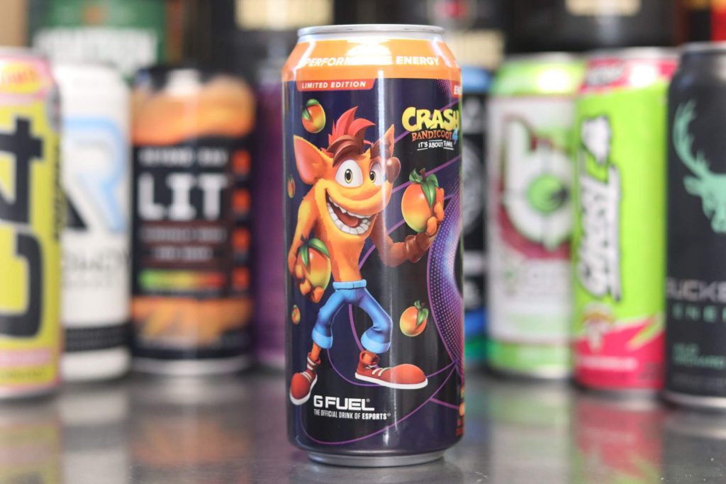 closeup of g fuel energy drink with crash bandicoot cartoon wearing jeans and shoes holding exotic fruits in its hands while it smiles with background of 8 other energy drink brands