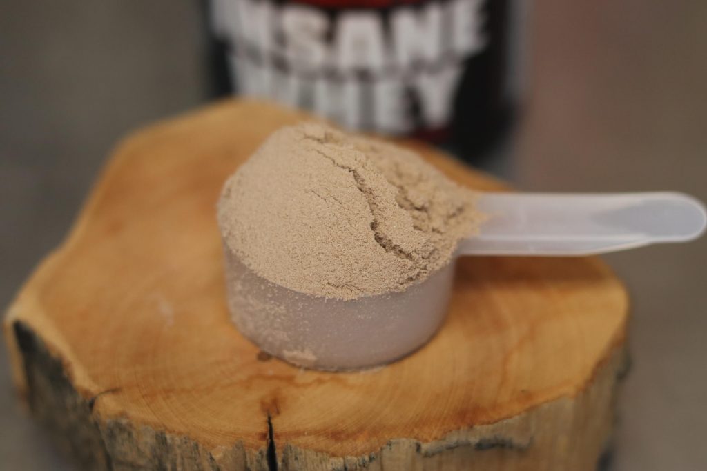 closeup of scoop of insane labz insane whey chocolate flavor on a wooden block with a small canister of insane whey behind it and blurred out
