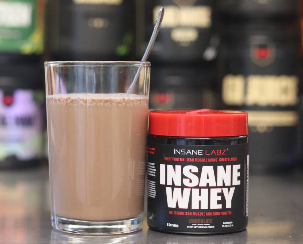 glass with spoon sticking out filled with brown mixture of Insane Labs Insane Whey and water alongside a small black tub of insane whey with a red lid