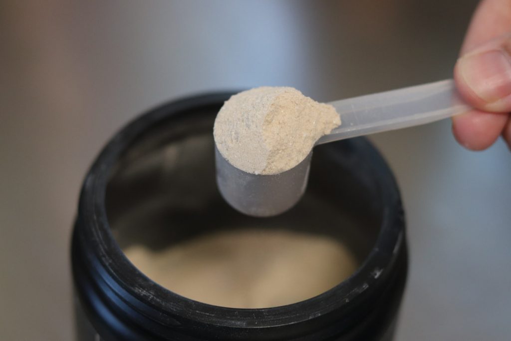 closeup of scoop of redcon1 double tap powder being held by two fingers with a black tub of double tap powder in the background