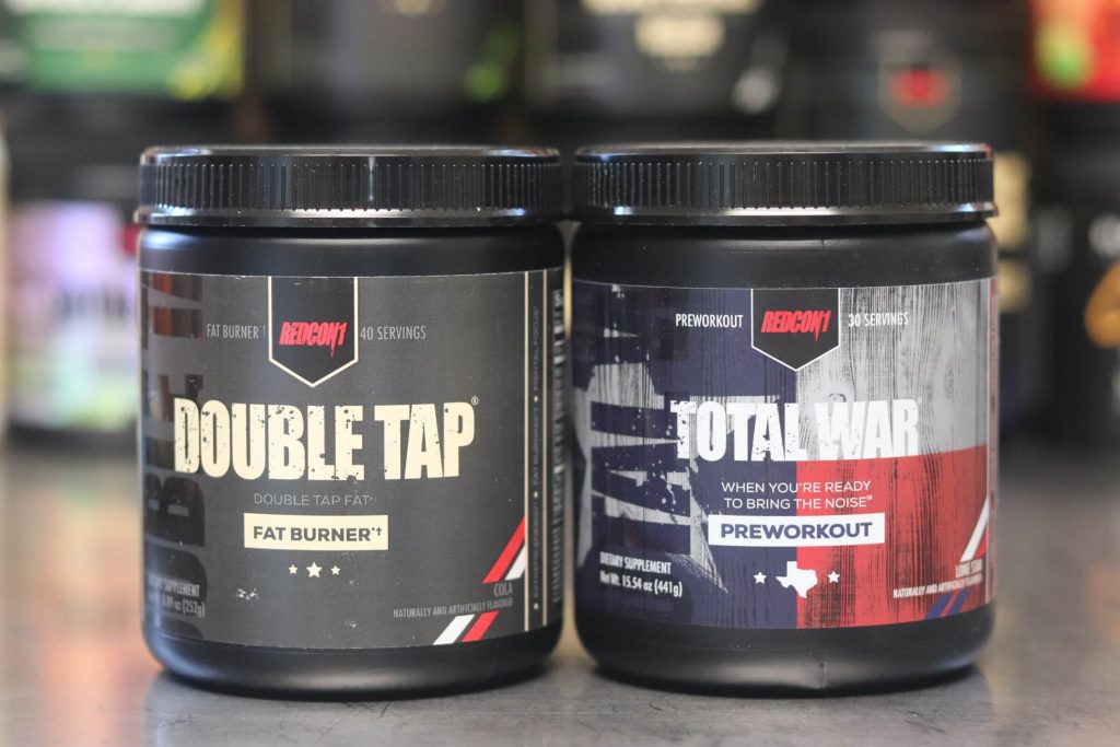 black tub of redcon1 double tap fat burner powder in cola flavor with black label next to black tub of total war with red white and blue label in lone star flavor