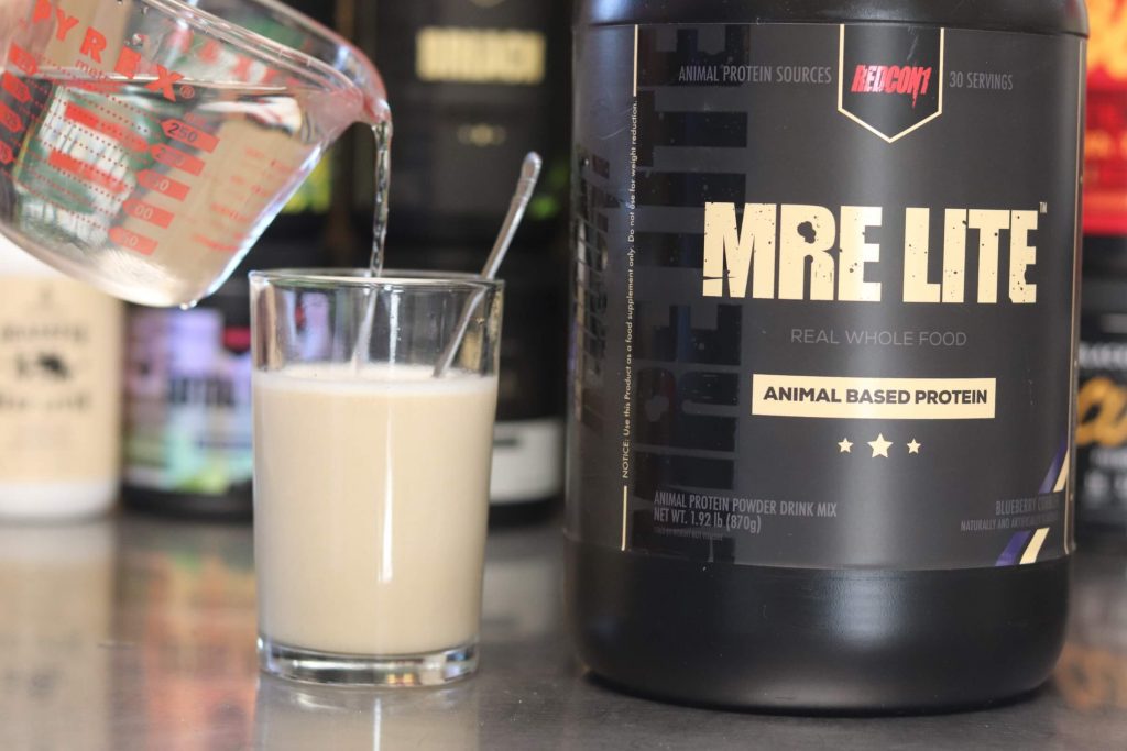 Pyrex glass pouring water into glass mixed with Redcon1 MRE Lite protein powder next to a giant black tub of MRE Lite protein powder