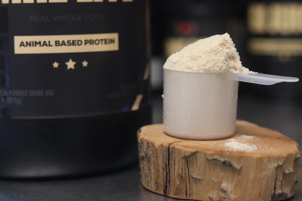 Closeup of heaping scoop of Redcon1 MRE Lite protein powder sitting on a wooden block next to a black tub of the MRE Lite protein powder